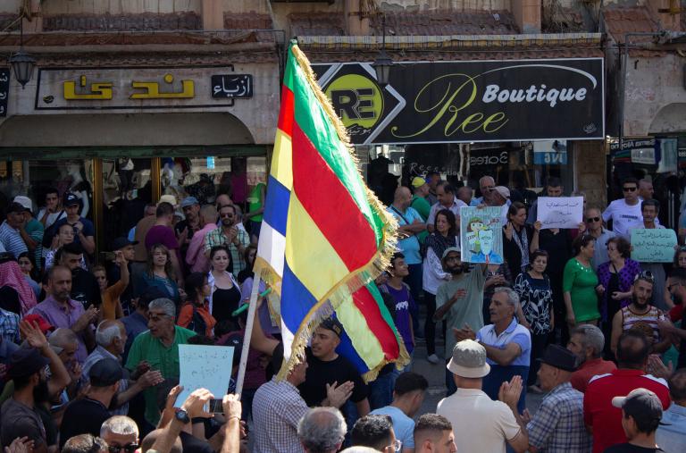 Protesting Syrians in the town of Suwayda hold signs and Druze flags - source: Reuters